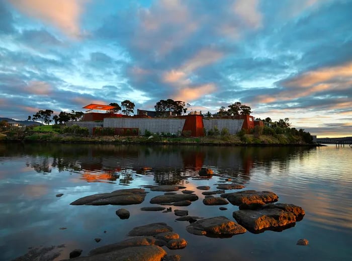 The MONA museum’s buildings rise from an islet at sunset, surrounded by water, with small stones peeking above the surface and fluffy clouds drifting across a blue sky.