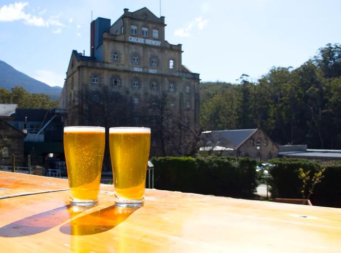 Two full glasses of beer rest on a table, with the majestic stone facade of Cascade Brewery rising in the background.