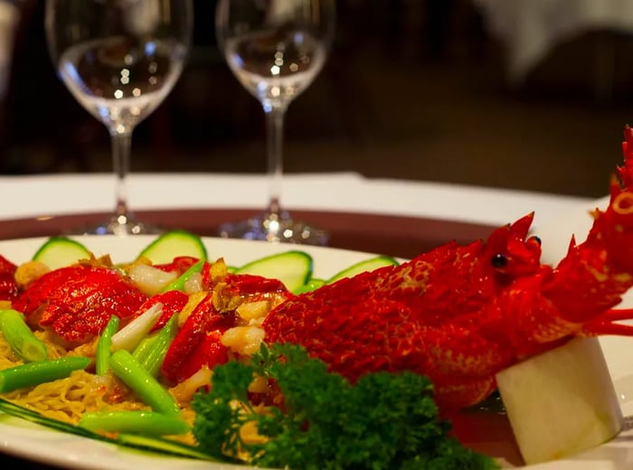 A long platter showcases a whole cooked lobster adorned with noodles and sliced vegetables, elegantly presented on a white tablecloth, with several wine glasses in the background.