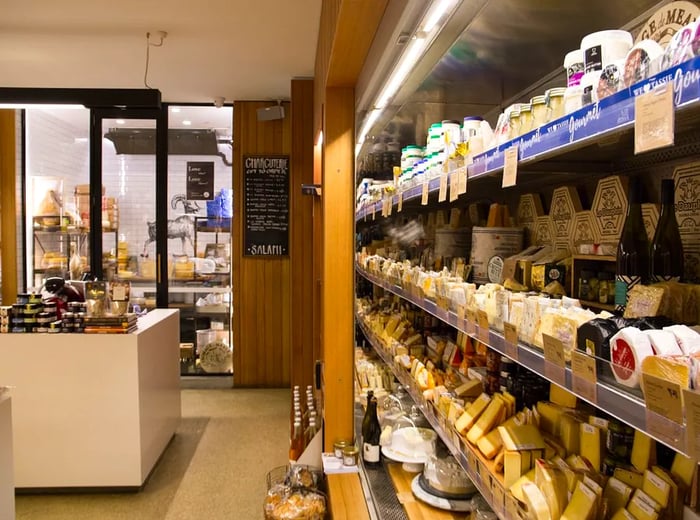 On the right, a cheese fridge filled with various cheeses and their labels, with a cold storage room and a counter ahead displaying a selection of jams and spreads for sale.