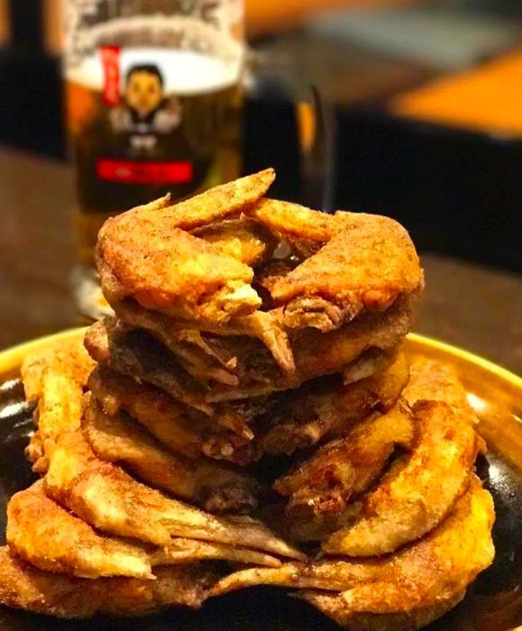 A towering stack of fried chicken wings, generously sprinkled with spices, rests on a ceramic plate atop a wooden table, with a blurred beer mug featuring a cartoon mascot in the background.