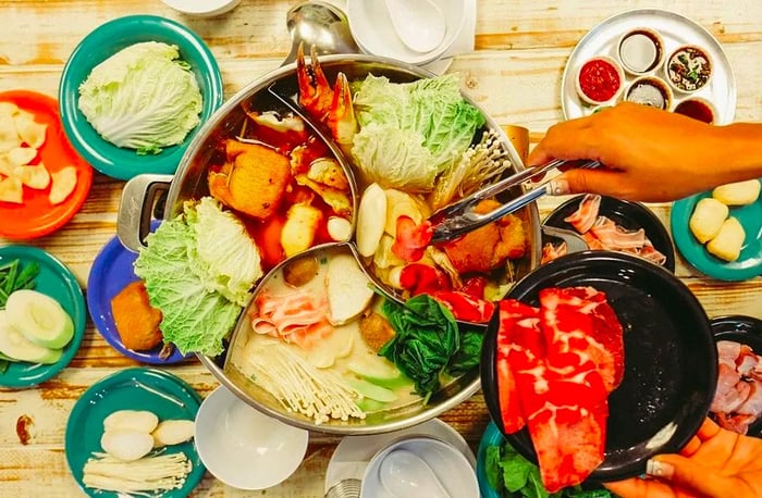 From above, a divided hot pot brimming with vegetables, seafood, and meats, while a hand adds more ingredients using tongs, with various condiments and ingredients spread across the table.