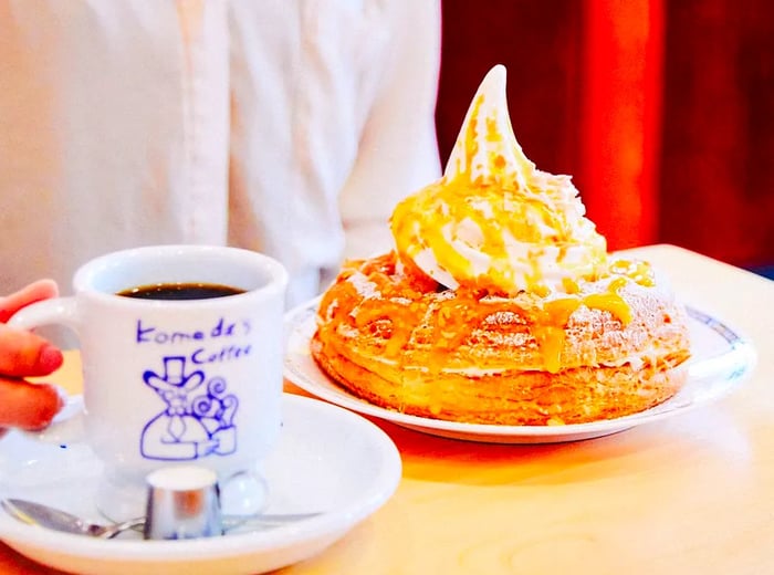 A customer enjoys coffee at a small table, holding a branded mug, alongside a large pastry topped with a swirl of soft serve ice cream.