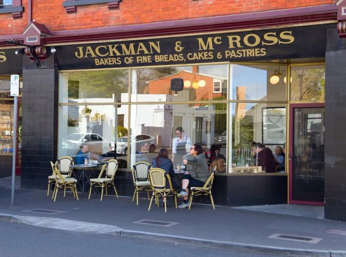 The vintage sign for Jackman & McRoss adorns a red brick building, showcasing large windows into the dining area, where guests are seated at outdoor patio tables.