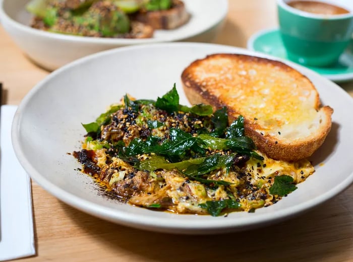 A shallow bowl brimming with scrambled eggs mixed with tender duck, enhanced by curry leaves and lemongrass, accompanied by a thick slice of sourdough, with other dishes softly blurred in the background.