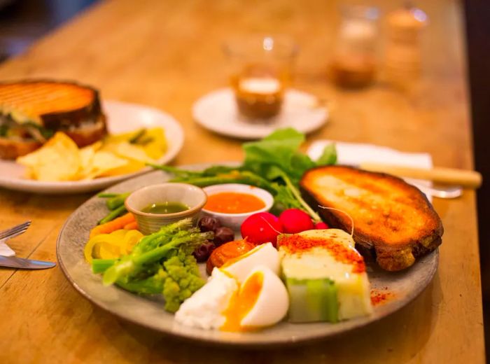 A beautifully arranged plate featuring a variety of colorful vegetables, a soft-boiled egg sliced open, a thick slice of toast, and several small bowls of dipping sauces.