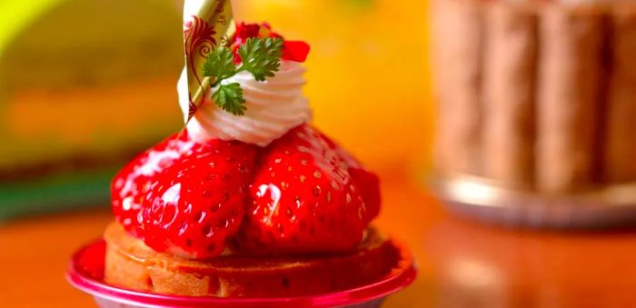 A close-up view of glossy strawberries atop a small cake adorned with whipped cream and a sprig of herb.