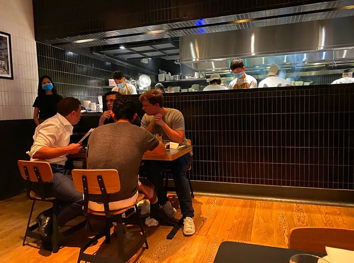 A group of four men sits at a table in front of a kitchen line where everyone is wearing masks.