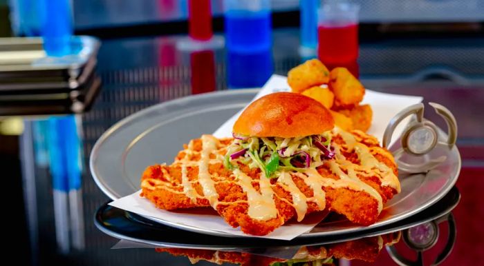 A sandwich featuring a massive fried chicken cutlet on a small bun, topped with slaw and sauces, alongside a generous serving of tater tots on a metal tray.