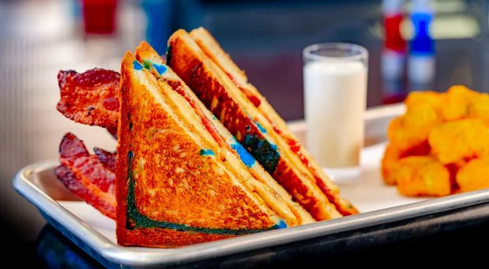 A bacon peanut butter and jelly sandwich on blue-streaked bread, cut into triangles, sits on a metal tray next to tater tots and a smoothie shooter.