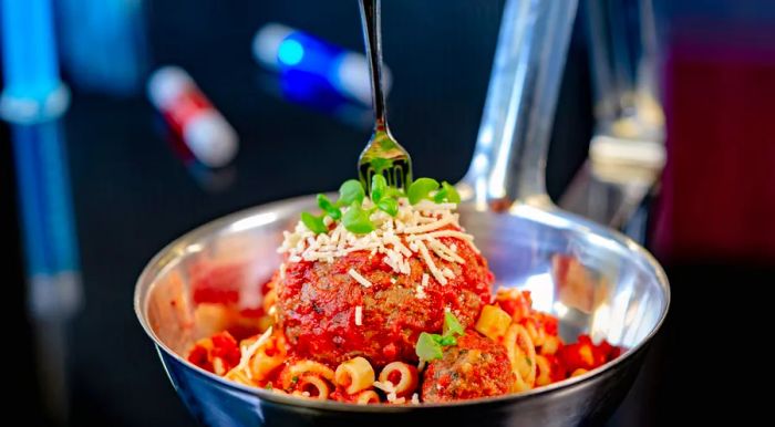 An enormous meatball resting on a bed of pasta, drizzled with marinara sauce, topped with grated cheese, herbs, and a tiny plastic fork stuck in the meatball.