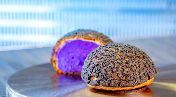 A sizable cream puff filled with purple cream and topped with a layer of black baked sugar, displayed on a metal table.