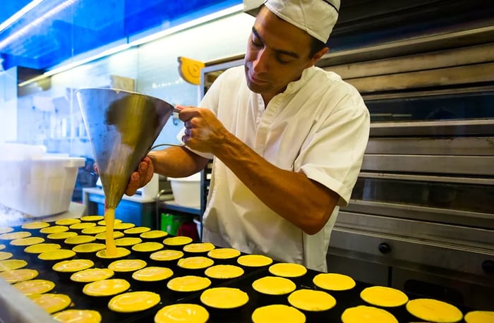 A baker carefully fills a row of delicate egg tarts.