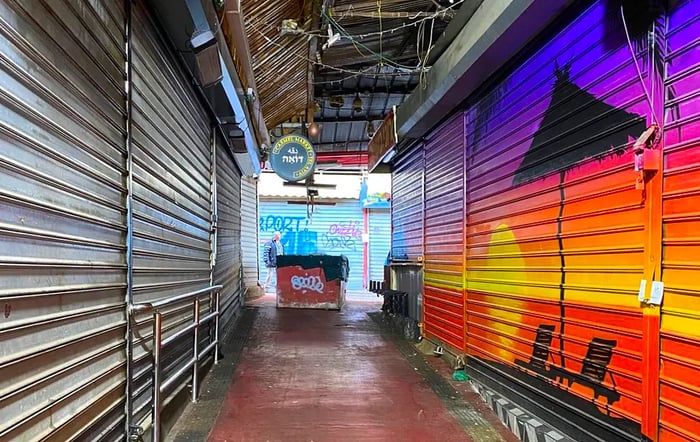 The empty stalls of the Carmel market are closed, their metal gates down, adorned with painted scenes of sunsets.