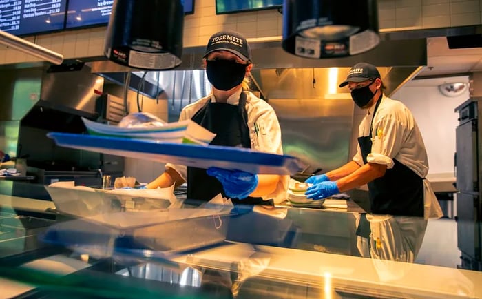 A woman stands behind the restaurant counter wearing a mask and a cap that reads 'Yosemite.'