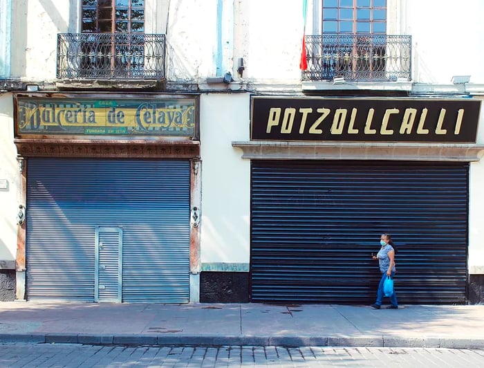 A woman in a mask walks by closed shops, carrying a bag.