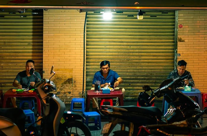 Three men enjoy their meals at small plastic tables set up in front of motorcycles.