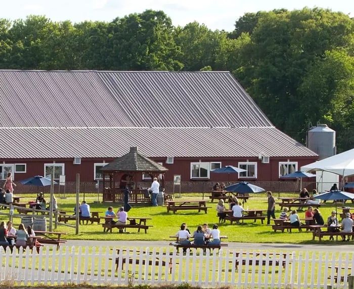 Tables set under umbrellas are positioned outside a barn, maintaining several feet of distance between them.