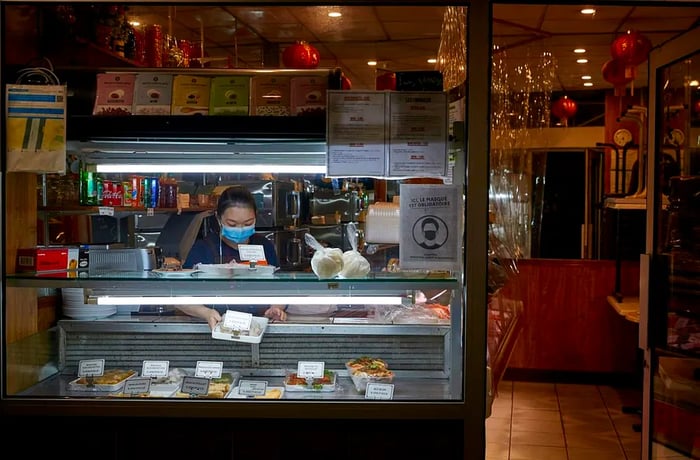 A woman wearing a mask leans over a deli case at night.