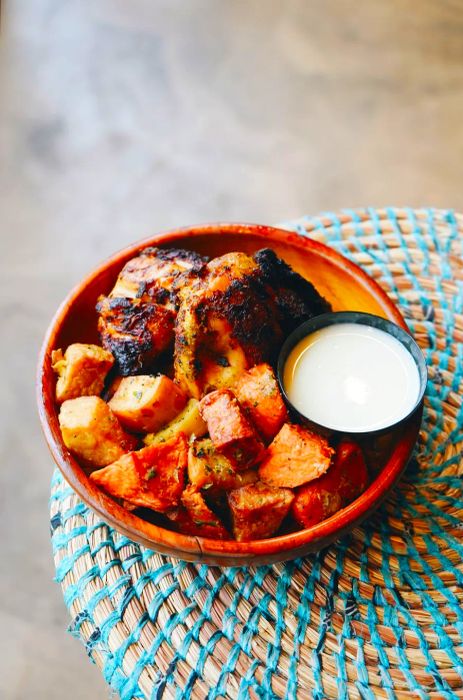 A wooden bowl showcases a serving of braised chicken paired with sweet potatoes, accompanied by a small container of white sauce.