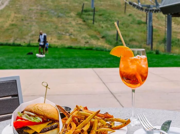 A portrait captures a vast green mountain slope in summer, with a platter of burgers and fries alongside an Aperol spritz cocktail topped with orange in the foreground.