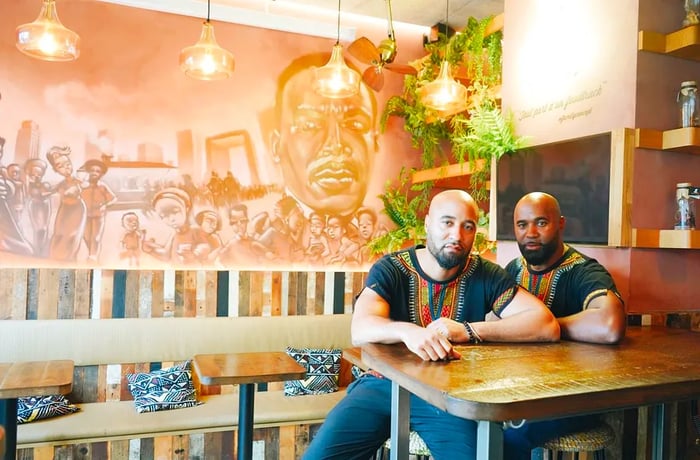 Rudy and Joël Lainé are seated at a table in the dining area of New Soul Food-Le Maquis. Behind them is a large mural, complemented by small tables in front of a banquette adorned with pillows.