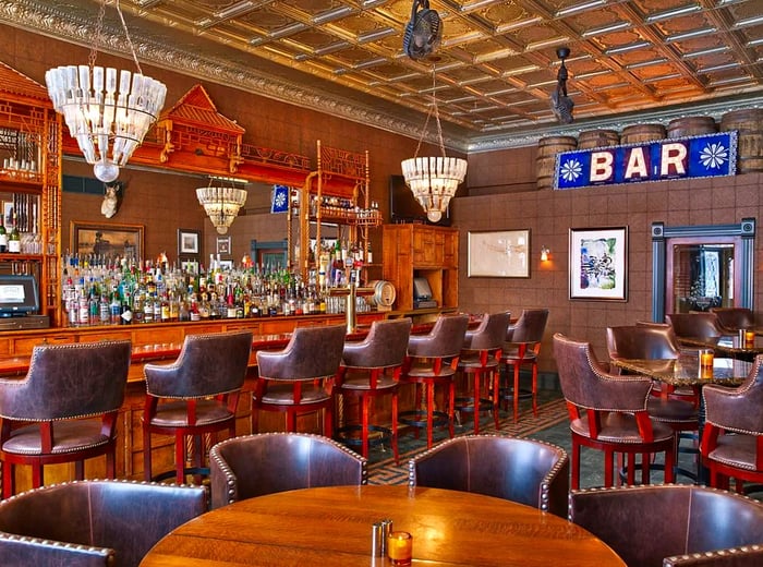 An interior view of a bar featuring high wooden tables, leather-upholstered stools, elegant chandeliers hanging above the bar, and a large carved wooden mirror adorning the back wall.