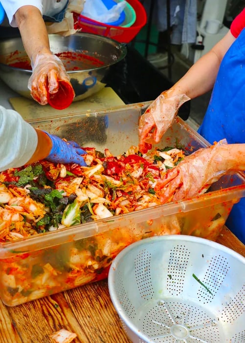 Gloved hands combine kimchi in a large plastic container