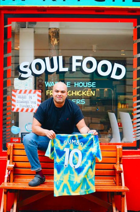 Lionel Chauvel-Maga sits on a wooden bench outside his restaurant, holding a blue tie-dye t-shirt emblazoned with “Gumbo 10.” Above him, the words “Soul Food” are displayed in white letters on the restaurant's window.