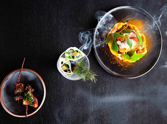 From above, three small dishes: a pair of oysters with aromatic herbs, skewers, and a seafood plate.