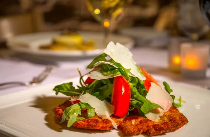 A plate featuring a salad of lettuce, tomato, and cheese atop breaded chicken, with a softly blurred table set in the background.