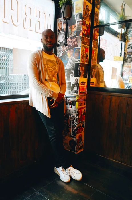 Brice Naranassamy stands inside Mama Jackson, facing away from a window. Beside him is a wall adorned with a collage of photos, posters, and magazine covers, along with a mirror reflecting his profile.