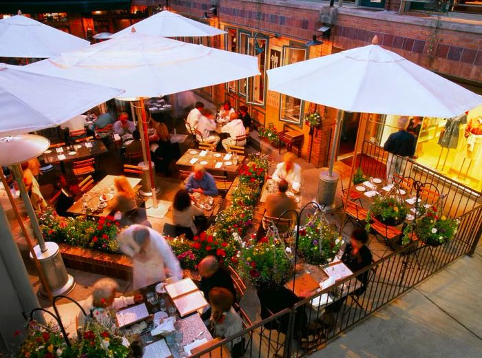 An aerial view of a lively patio filled with tables of patrons, servers bustling about, and white umbrellas providing shade.