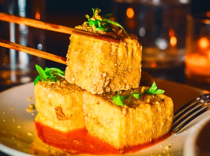 A vertical shot captures a pair of chopsticks reaching for a piece of crispy tofu from a plate featuring three pieces of salt-and-pepper tofu.