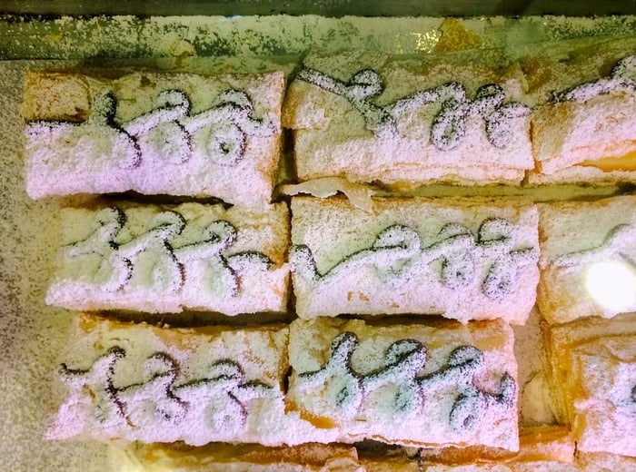 A bird's-eye view of a display case filled with snow puffs (rectangular layered delights adorned with powdered sugar and drizzled with chocolate ganache)