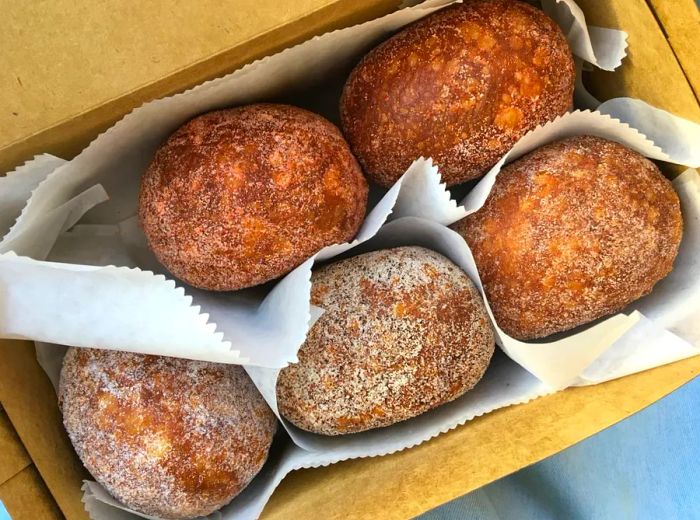 An overhead view of an open cardboard box showcasing five dark, sugar-coated doughnuts, each separated by paper liners