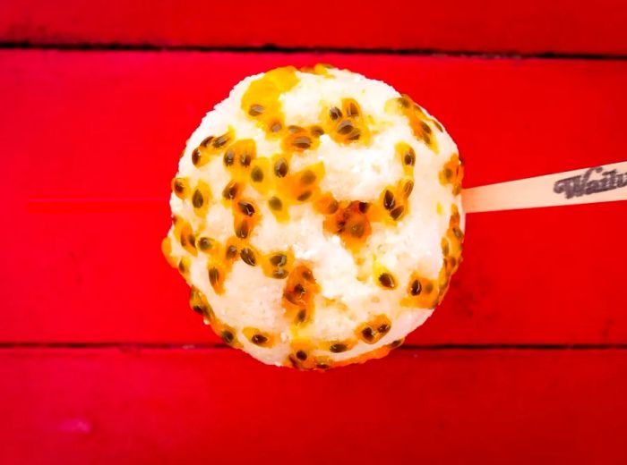 Aerial view of a cup of shave ice drizzled with syrup and sprinkled with fruit seeds, accompanied by a wooden spoon.