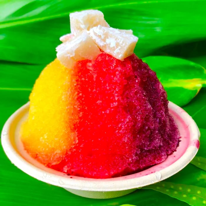 A vibrant mound of shave ice with an ombre effect, topped with small mochi pieces, set against a backdrop of tropical leaves.