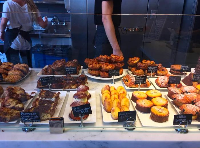 A display case filled with various pastries, each labeled individually, with staff working behind it