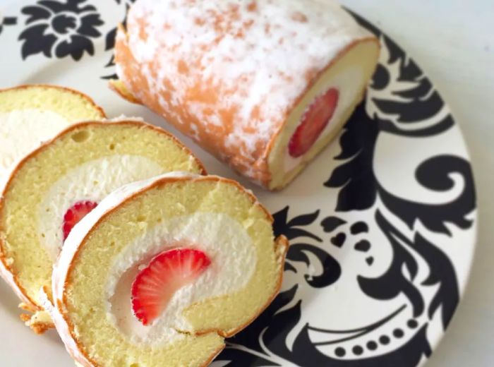 An aerial view of an artistic plate showcasing a half-rolled pastry dusted with sugar and filled with fruit at the center, accompanied by several slices displayed alongside to reveal the filling