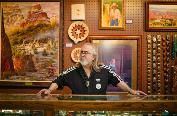 A man with a white beard and long hair, dressed in a black Adidas tracksuit, stands behind a glass counter showcasing Native American art and jewelry.