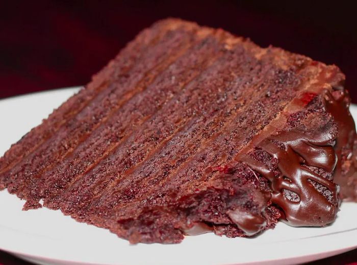 A slice of layered chocolate cake resting on a plate against a black backdrop