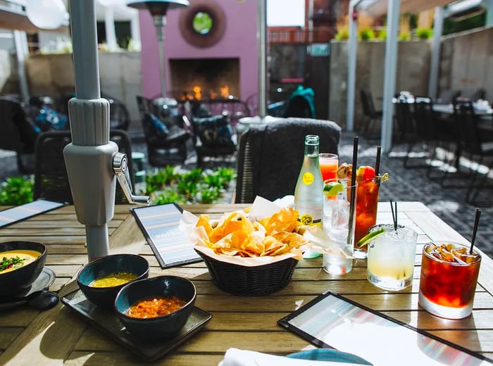 A table adorned with chips, an assortment of dips, and refreshing cocktails, set against a backdrop of a courtyard and an outdoor oven