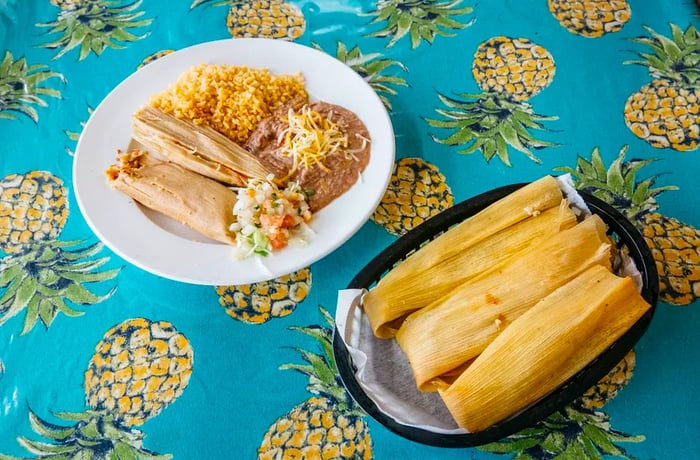 A white plate featuring a tamale accompanied by rice and beans rests beside a plastic basket containing three unwrapped tamales.