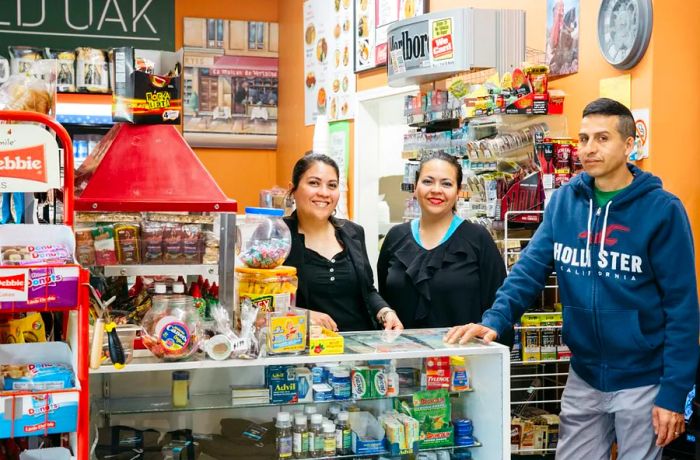 Two women are smiling from behind the counter of a convenience store, alongside a man who is also smiling in front of it.