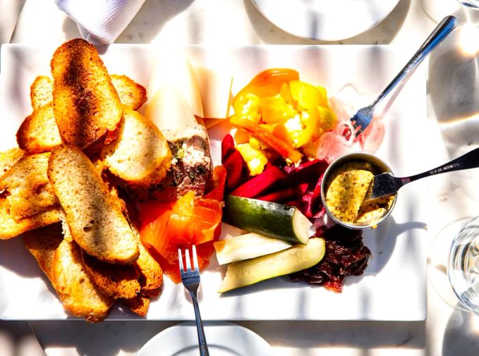 A beautifully arranged charcuterie board featuring pâté, sliced fish, pickled veggies, toasted bread, mustard, and an assortment of accompaniments