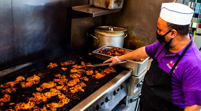 A man in a purple polo and chef’s hat operates a grill loaded with chicken pieces.