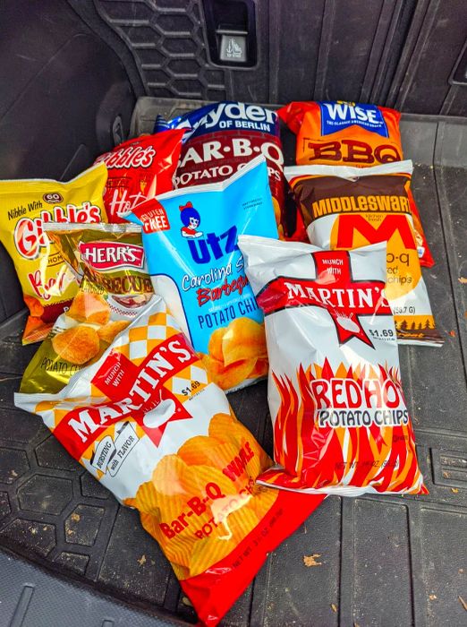 A collection of nine bags of potato chips, featuring options from Herr and Utz, stashed in the trunk of a car.