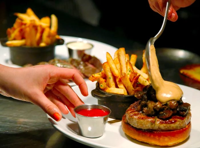 A chef adds a thick sauce to a burger, alongside a small dish of french fries