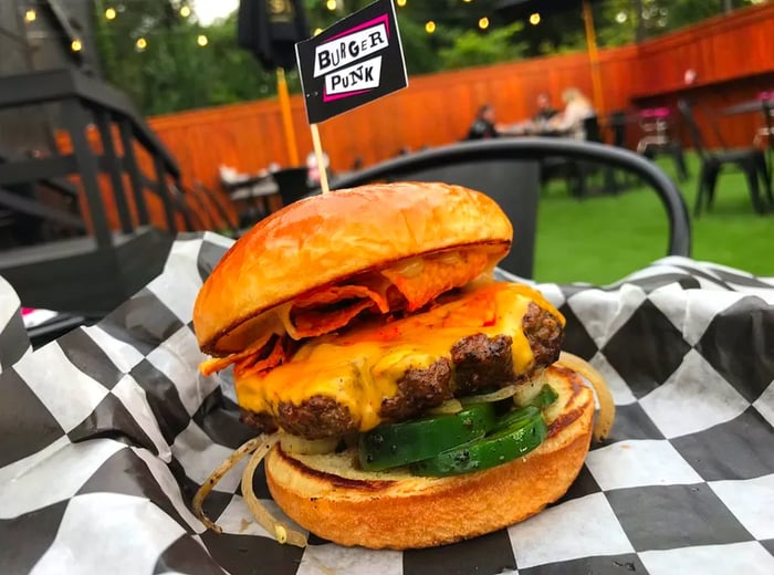 A cheeseburger adorned with Doritos chips resting on checkerboard wax paper, set against an astroturfed outdoor seating area